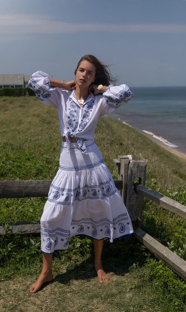 Front view of Seaside Blaire Tiered Skirt showcasing white linen tiered silhouette with blue paisley border print on a model at a grassy seaside cliff