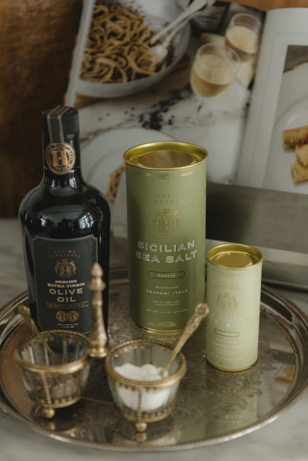 Front view of Full Range Bundle: dark olive oil bottle and green tins labeled Sicilian Sea Salt on a silver tray
