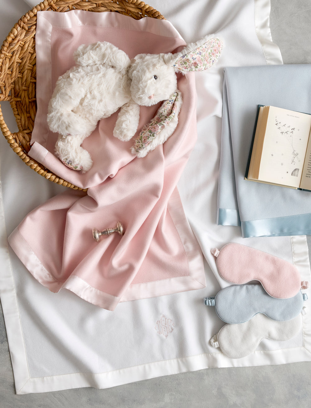 Three Dream Modal sleep masks in pink, blue, and cream arranged on white bedding beside a pink modal blanket, plush rabbit, and open book