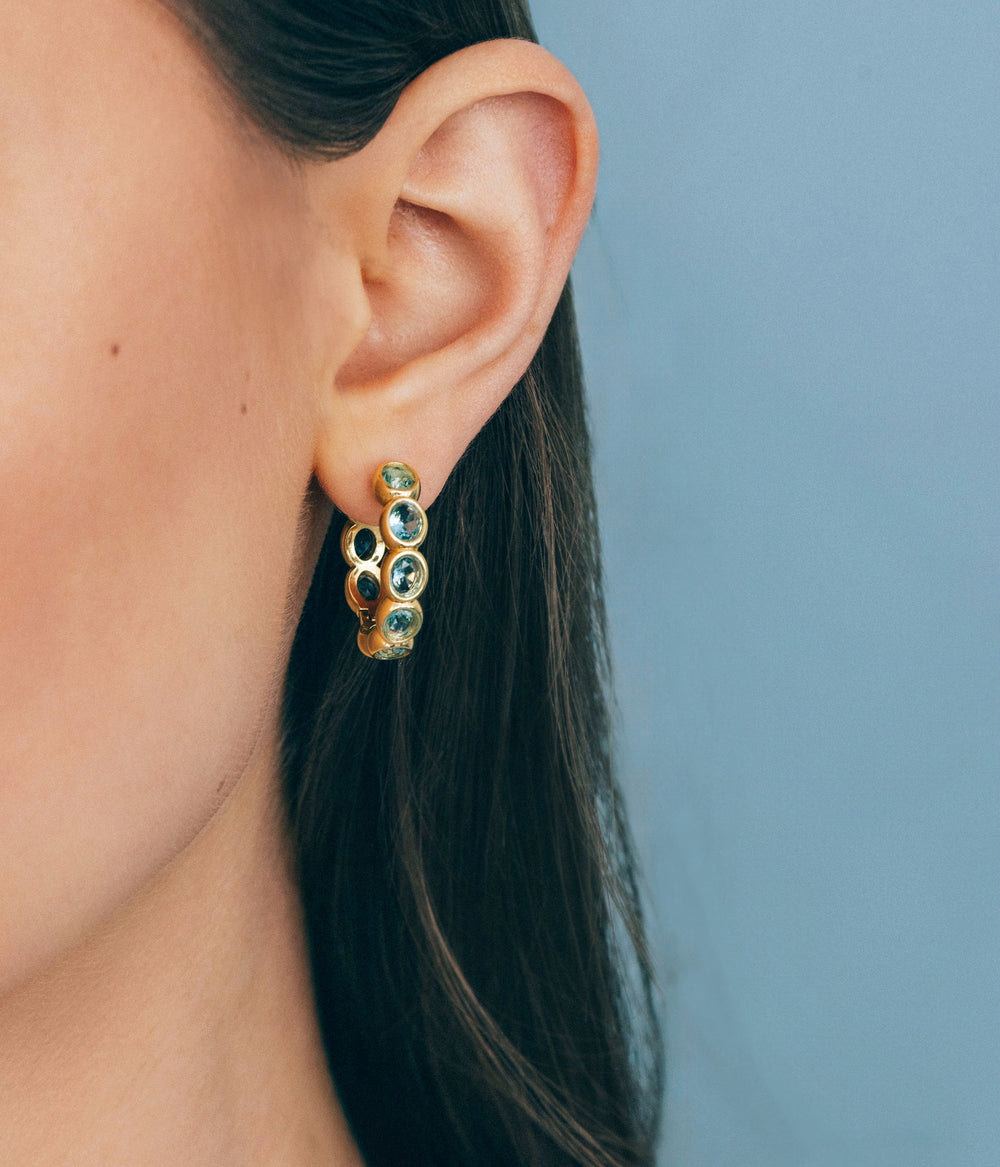 Close-up of Aurelia Hoop Earrings on model’s ear showcasing gold huggie silhouette with round aqua glass stones against pale blue background
