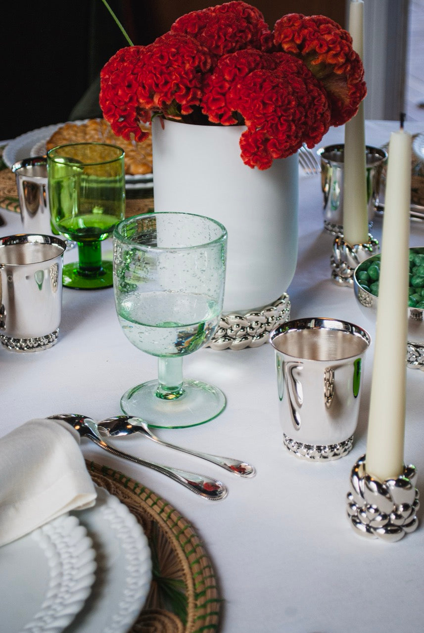 Silver-plated candlesticks with braided bases holding ivory taper candles on a white tablecloth beside green glassware and red floral centerpiece