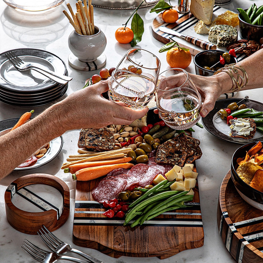 Two people clinking Amalia stemless white wine glasses over a charcuterie board with cheese, crackers, olives and fruit