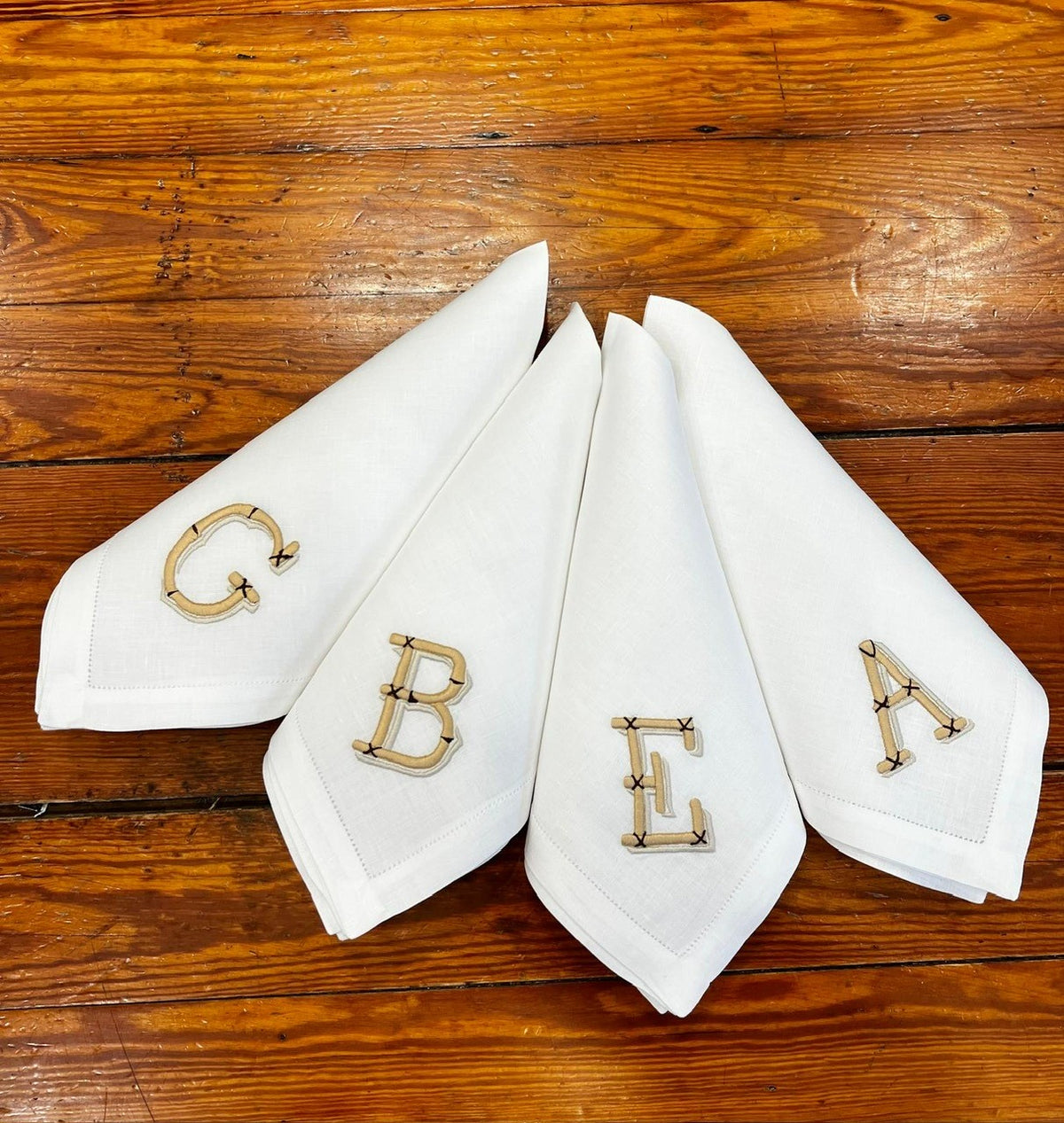 Four folded Bamboo Monogram linen napkins showing bamboo-inspired embroidered initials and hemstitch border on a wooden table