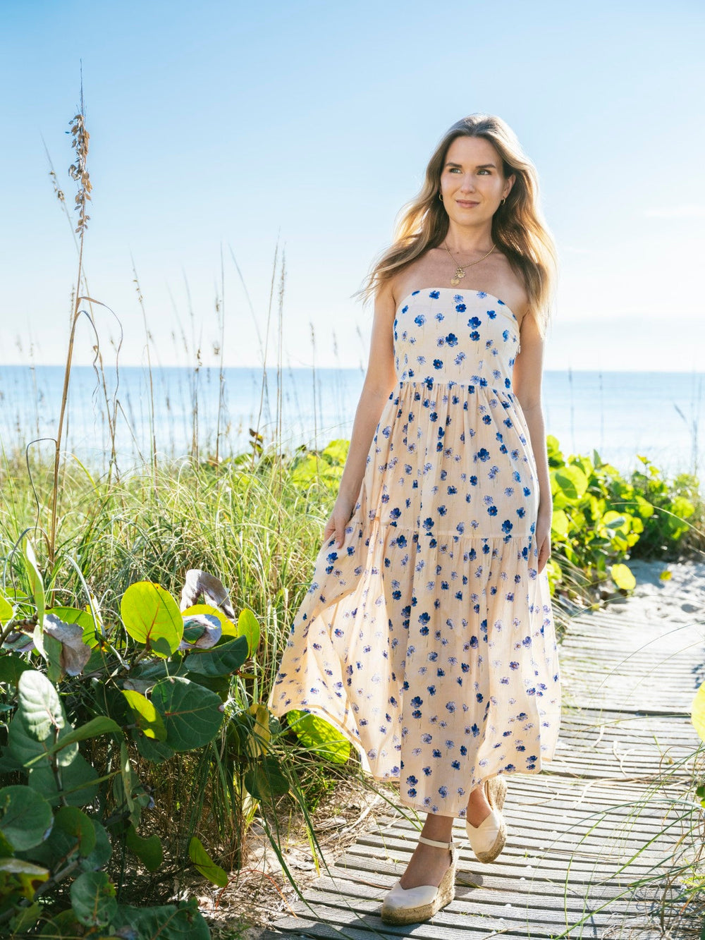 Front view of Barbie Dress in Bluebell, strapless with blue floral print on light peach, midcalf, on a beach boardwalk