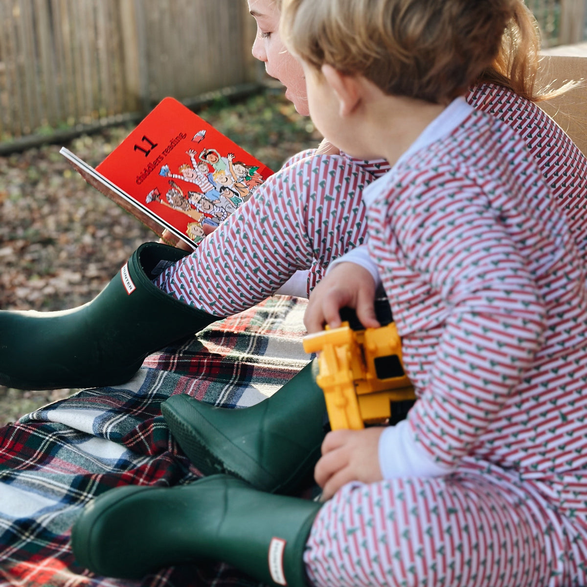 Two children in matching red-and-white Christmas tree stripe henley pajamas and green rain boots reading a picture book outdoors on a plaid blanket