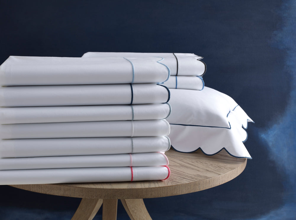 Front view of Butterfield bedding sheets showcasing scalloped edges and colored contrast piping on a round wooden table against a dark blue background