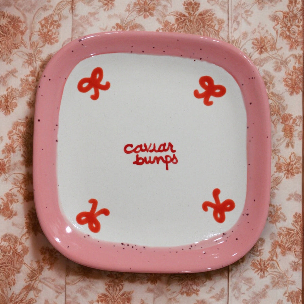 Top-down view of Retro “Caviar Bumps” tiny square plate showcasing a pink speckled rim, red handwritten “caviar bumps” center text and red corner motifs on a floral background