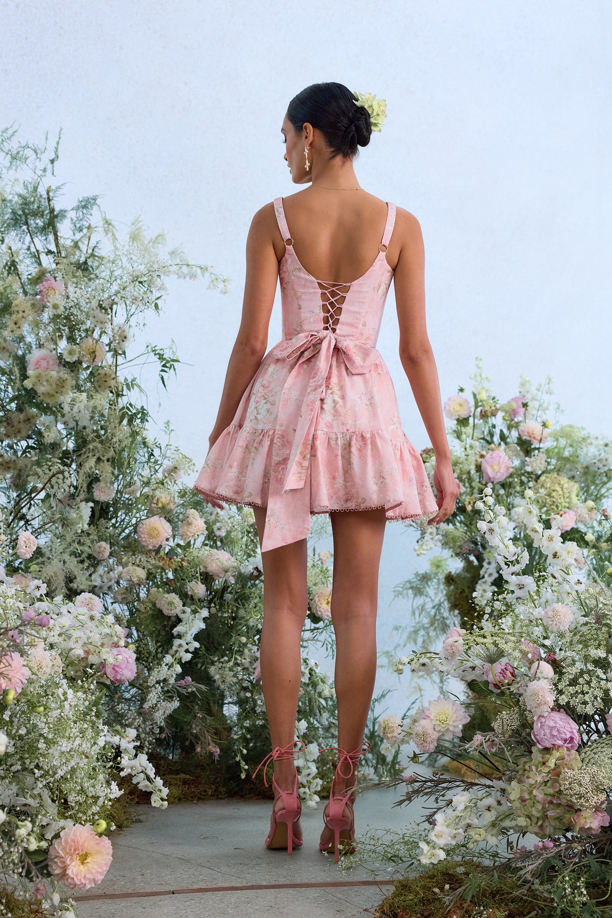 Back view of The Betina Dress in Light Pink Tapestry Rose showcasing lace-up back and bow detail with a flower-filled setting