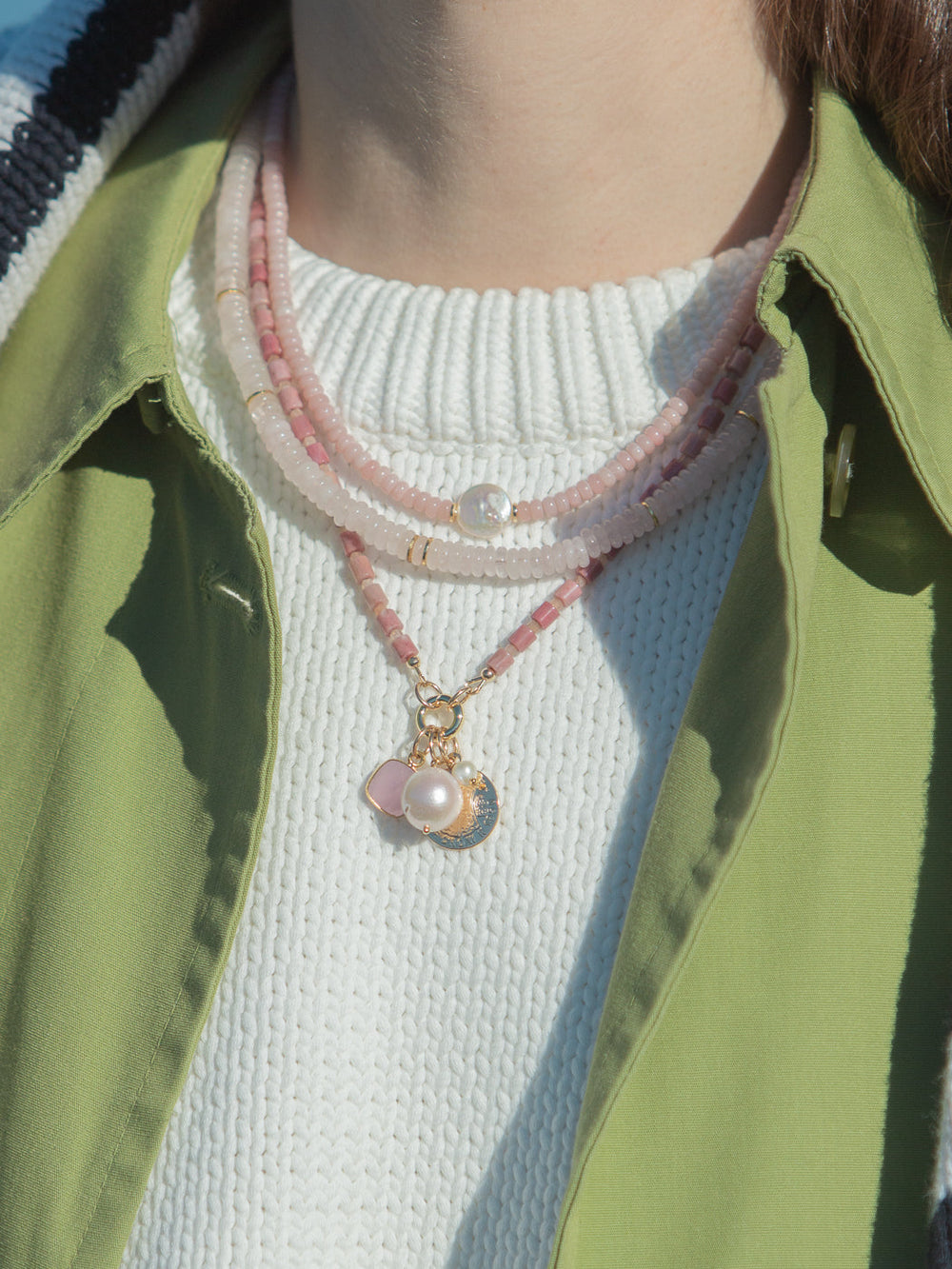 Close-up of Gretel Necklace in Pink with layered pink beads, freshwater pearl charm, and gold clasp on a white knit top