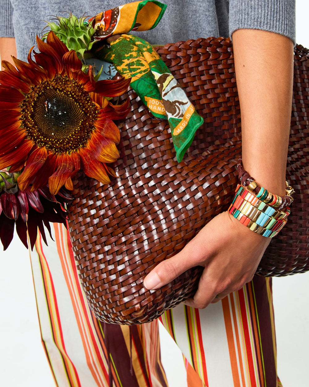 Close-up of wrist wearing Super Size Happy Cord Bracelet in brown showcasing gold-tone, pavé, and enamel slider charms with a woven brown tote background
