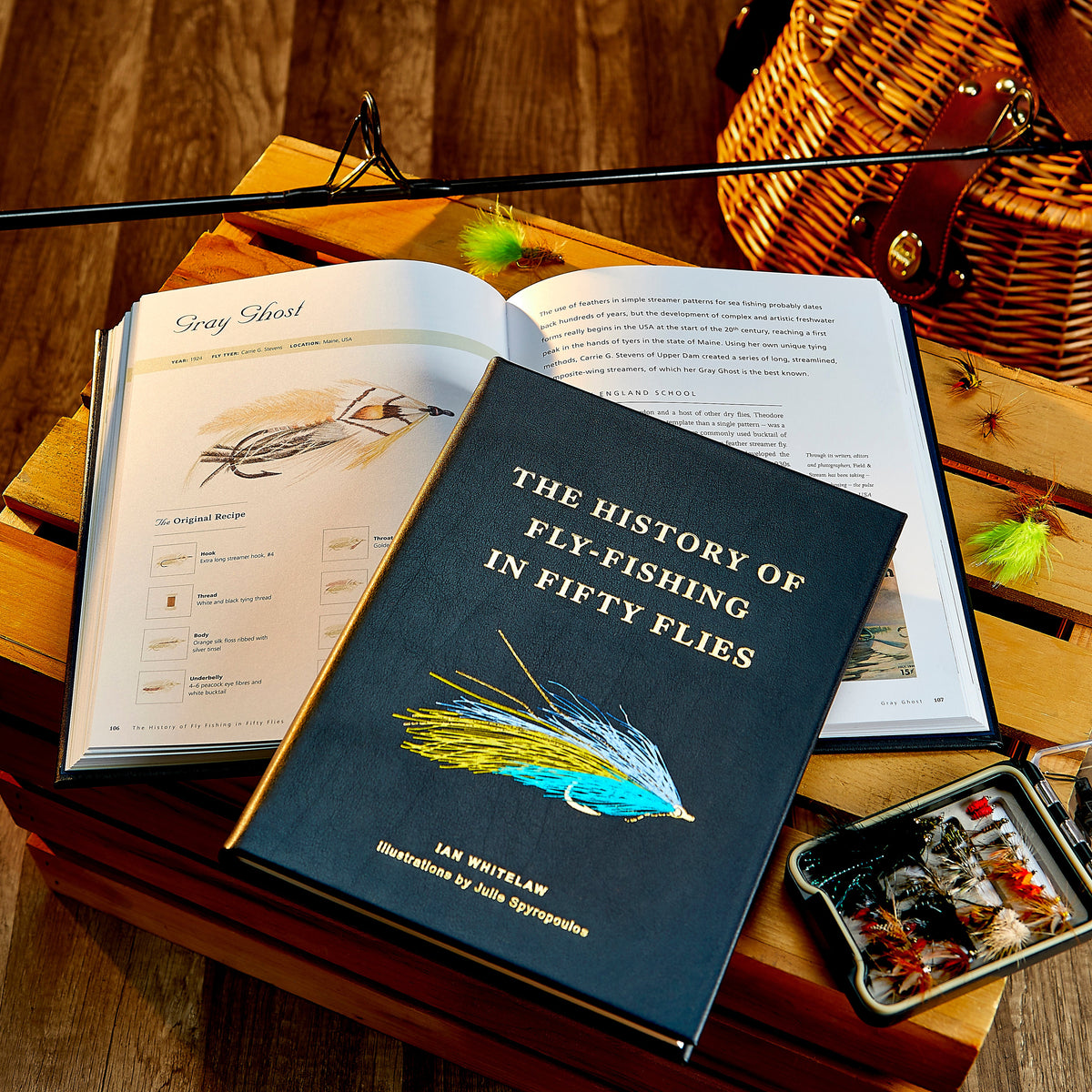 Open view of The History of Fly Fishing in Fifty Flies showcasing fly tying pages and fishing gear on a wood table