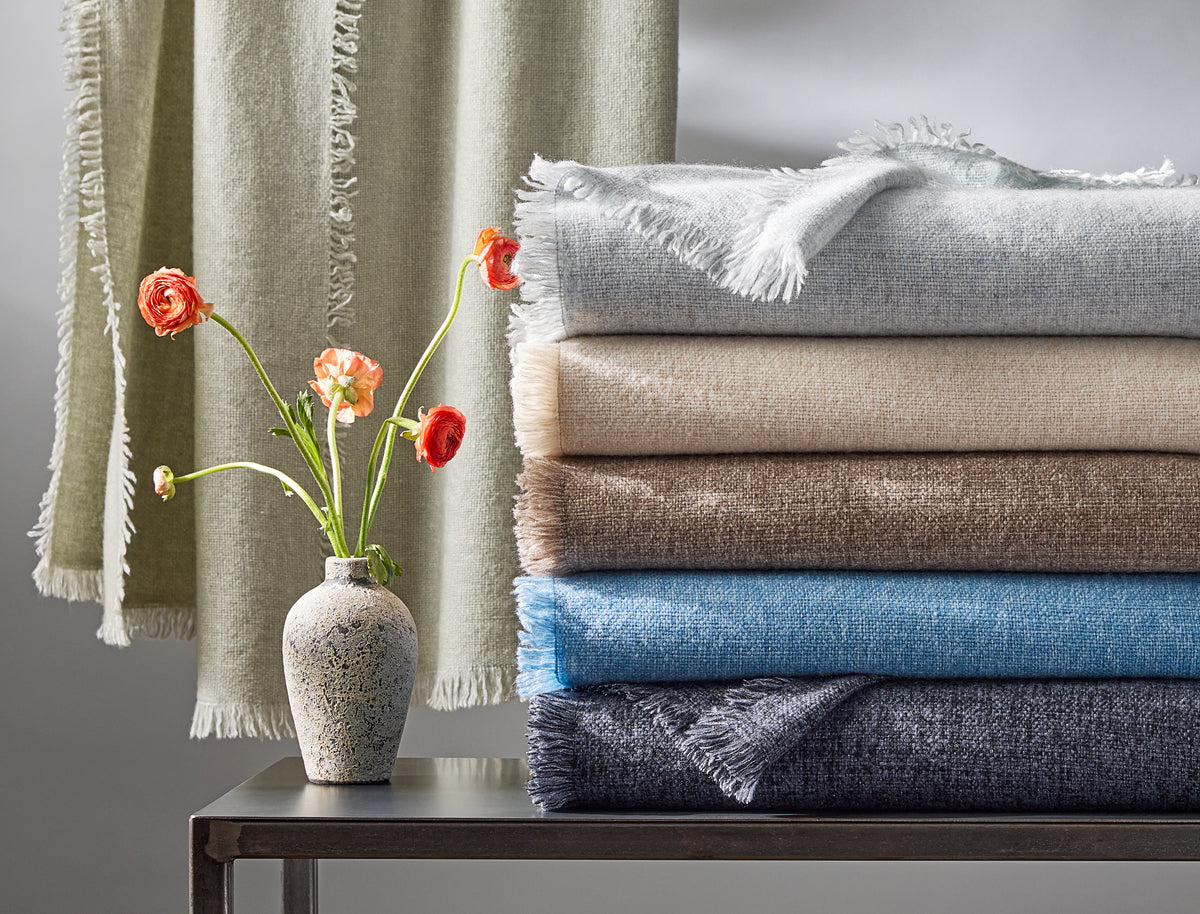 Side view of Maya throws stacked, showing fringe and soft cashmere blend, celadon throw hanging and vase with orange flowers