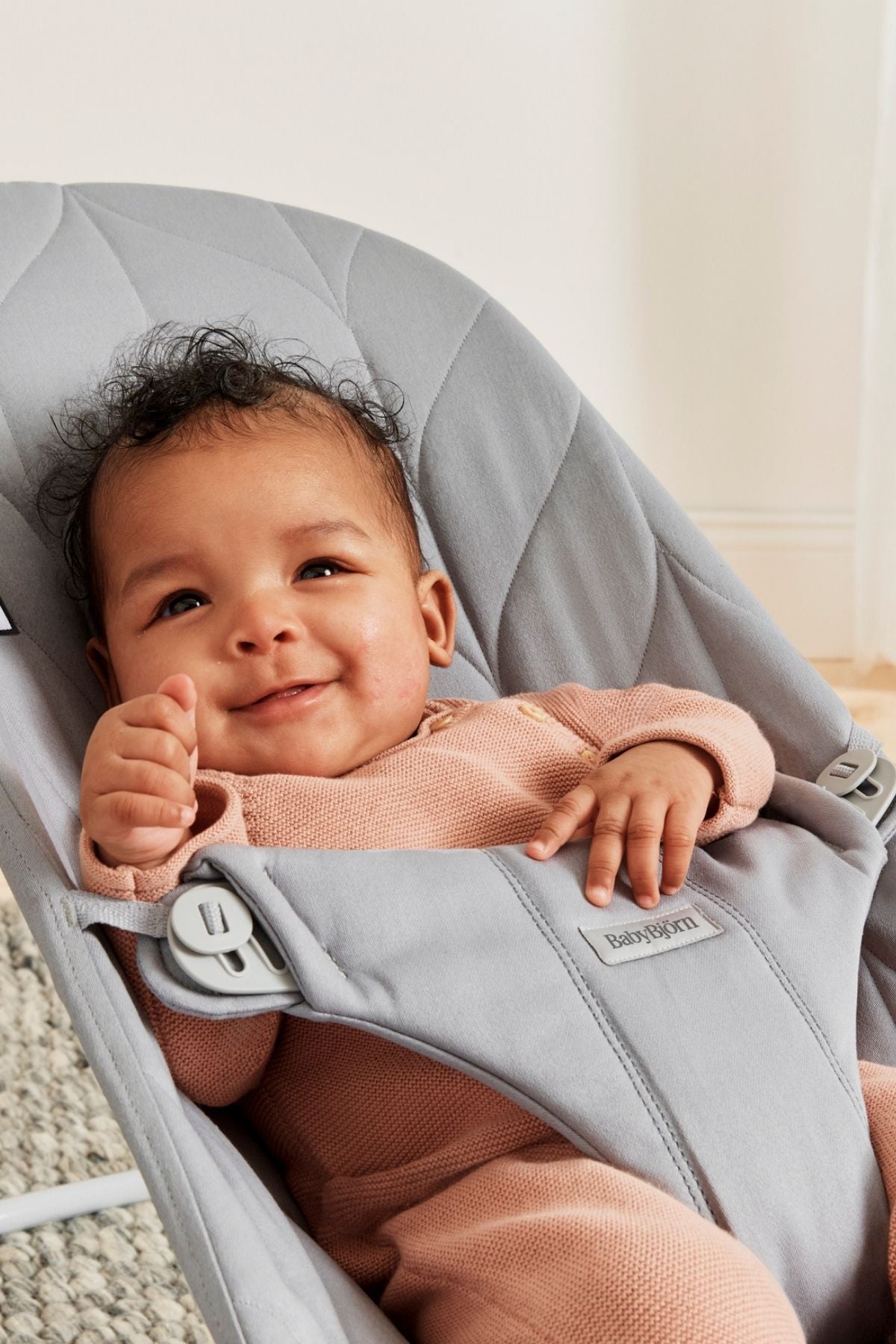 Front view of Bouncer Bliss in Petal Quilt Light Gray showcasing quilted seat and harness with a smiling baby in a neutral room