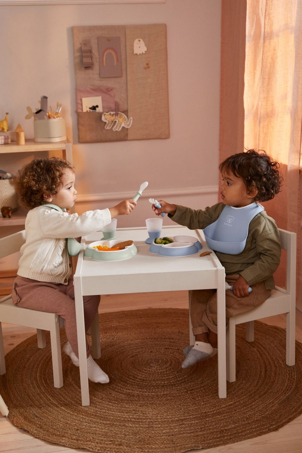 Two toddlers sitting at a small table sharing food using powder blue dinnerware pieces and bibs in a cozy playroom