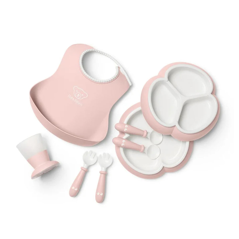 Overhead view of BabyBjorn 8-piece powder pink dinnerware set with trefoil divided plate, bib, cup, fork and spoon on white