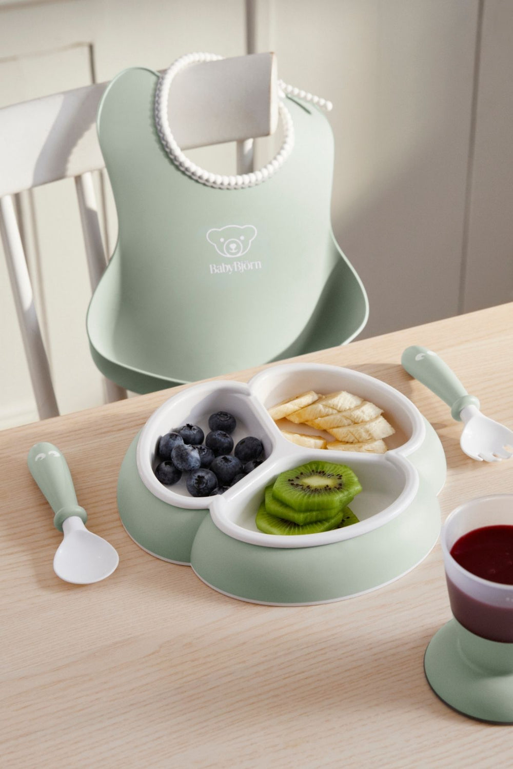 Angled top view of mealtime set in powder green showing trefoil divided plate, short-handled spoon and fork, and hanging bib