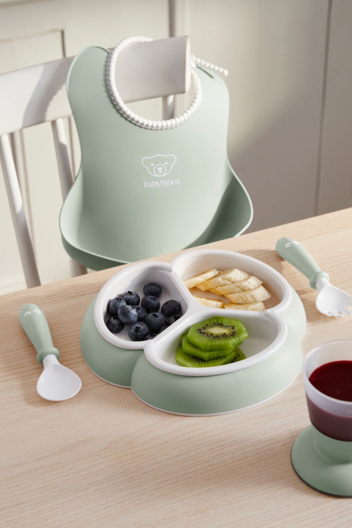 Angled top view of mealtime set in powder green showing trefoil divided plate, short-handled spoon and fork, and hanging bib