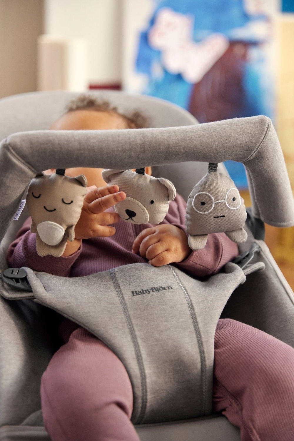 Close-up of Toy for Bouncer in Soft Friends Neutrals showing three neutral-toned hanging plush characters with stitched faces attached to a gray bouncer