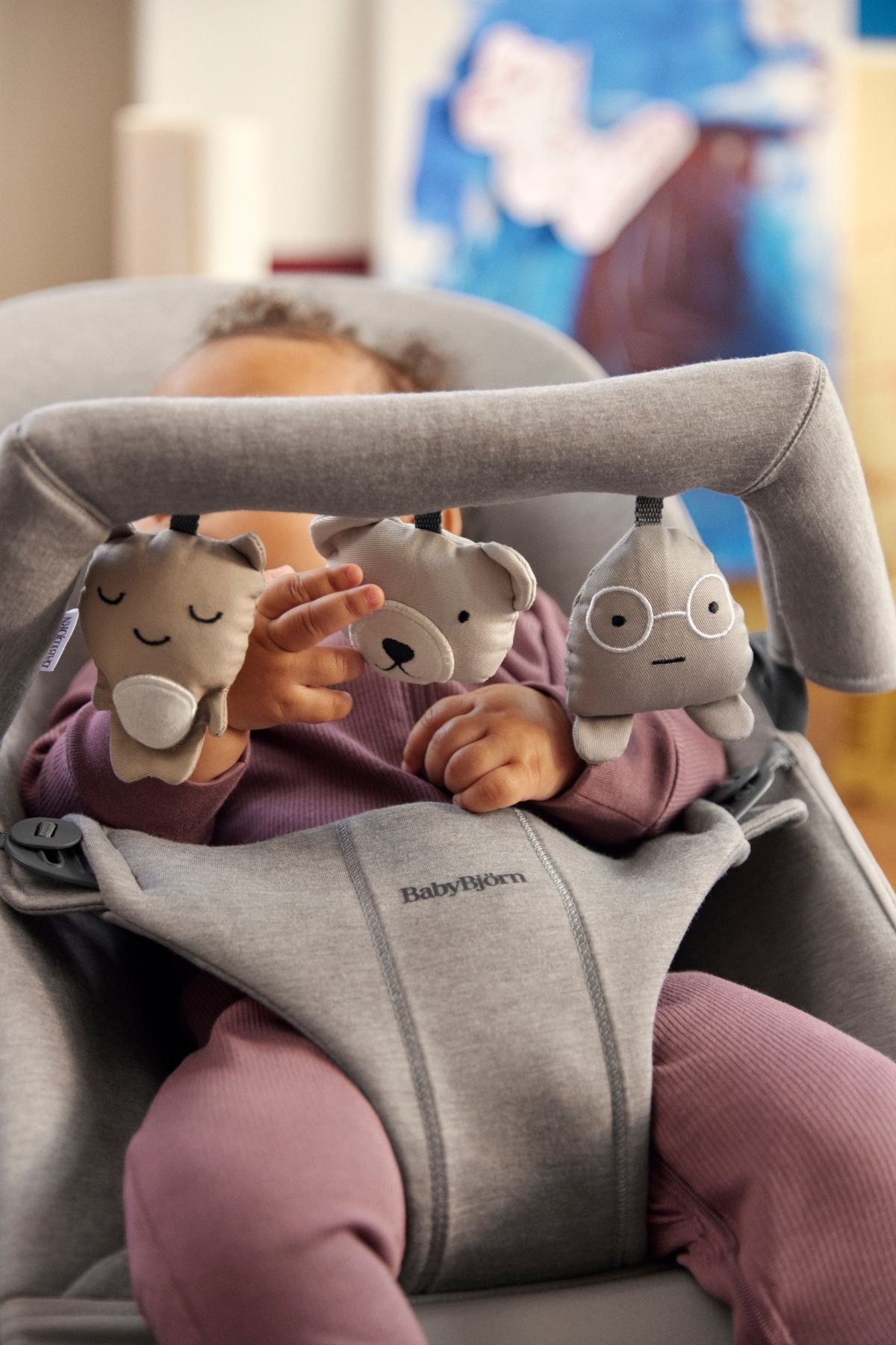Close-up of Toy for Bouncer in Soft Friends Neutrals showing three neutral-toned hanging plush characters with stitched faces attached to a gray bouncer