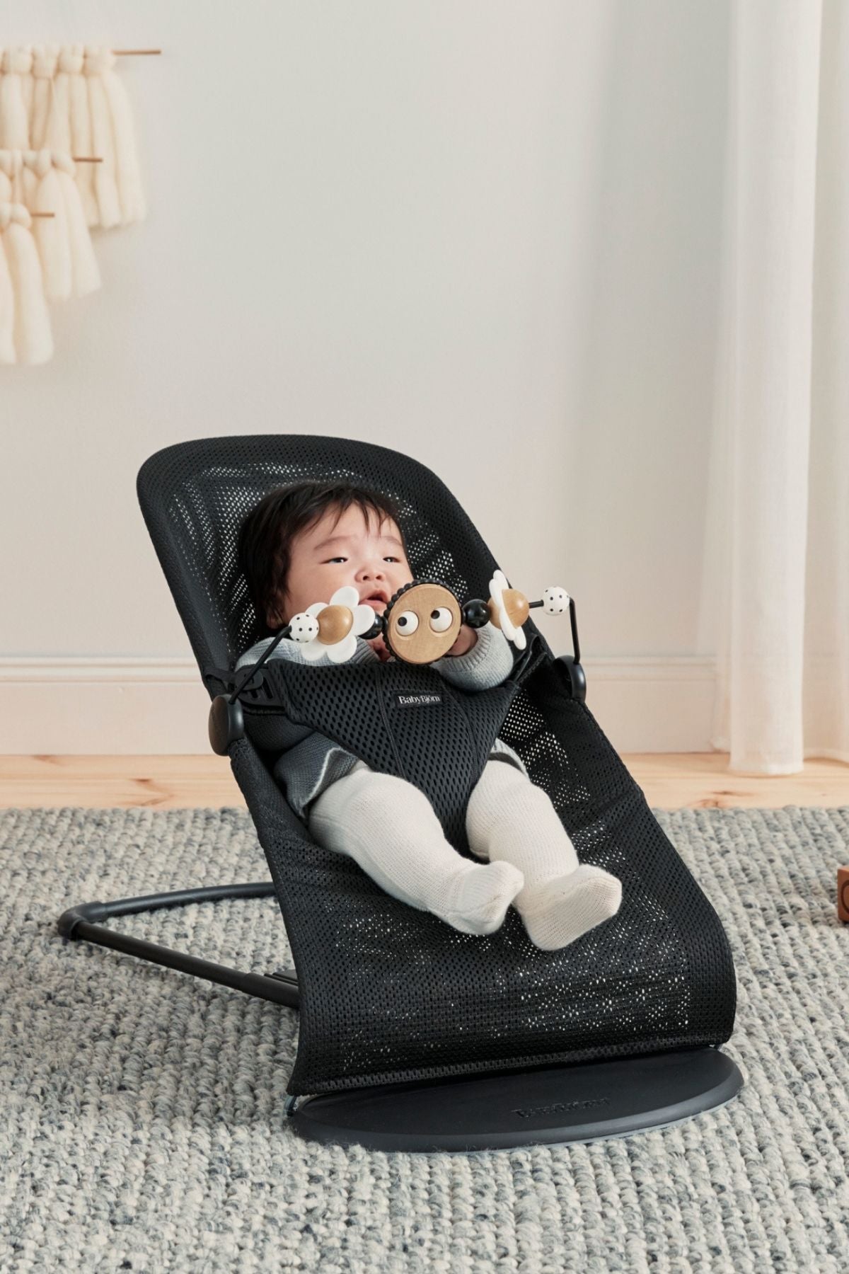 Front view of BabyBjörn Toy for Bouncer with black-and-white googly eyes and spinning wooden beads attached to a black bouncer in a nursery
