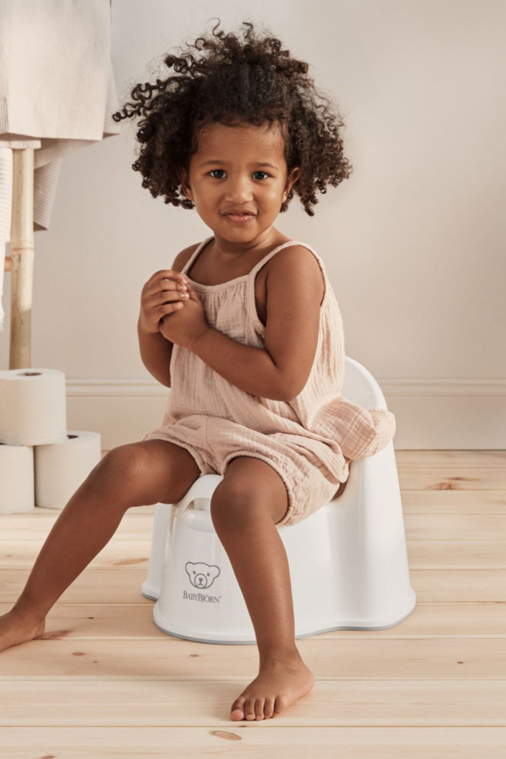 Front view of white BabyBjörn potty chair with ergonomic backrest and raised splash guard, toddler seated on light wood floor