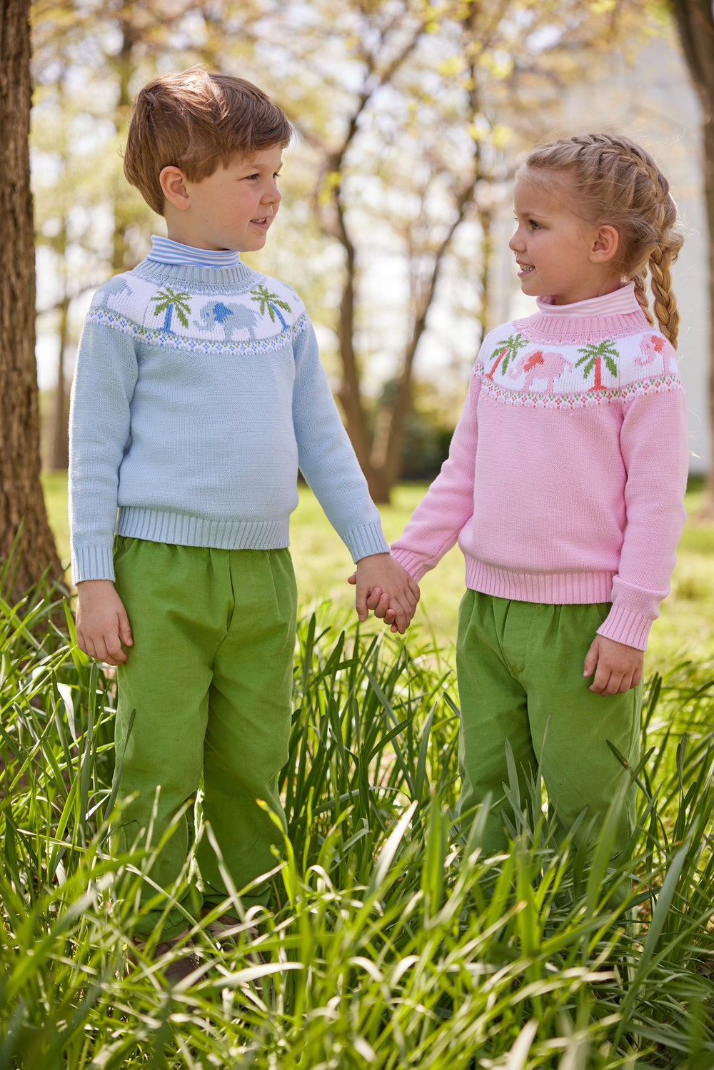 Two children holding hands outdoors, wearing light blue and pink striped turtlenecks layered under elephant yoke sweaters