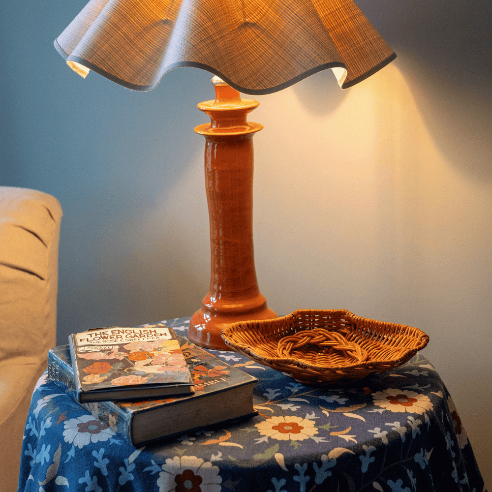 Angled view of Concha Dish showing scalloped shell shape and woven rattan on a floral side table with orange lamp and books