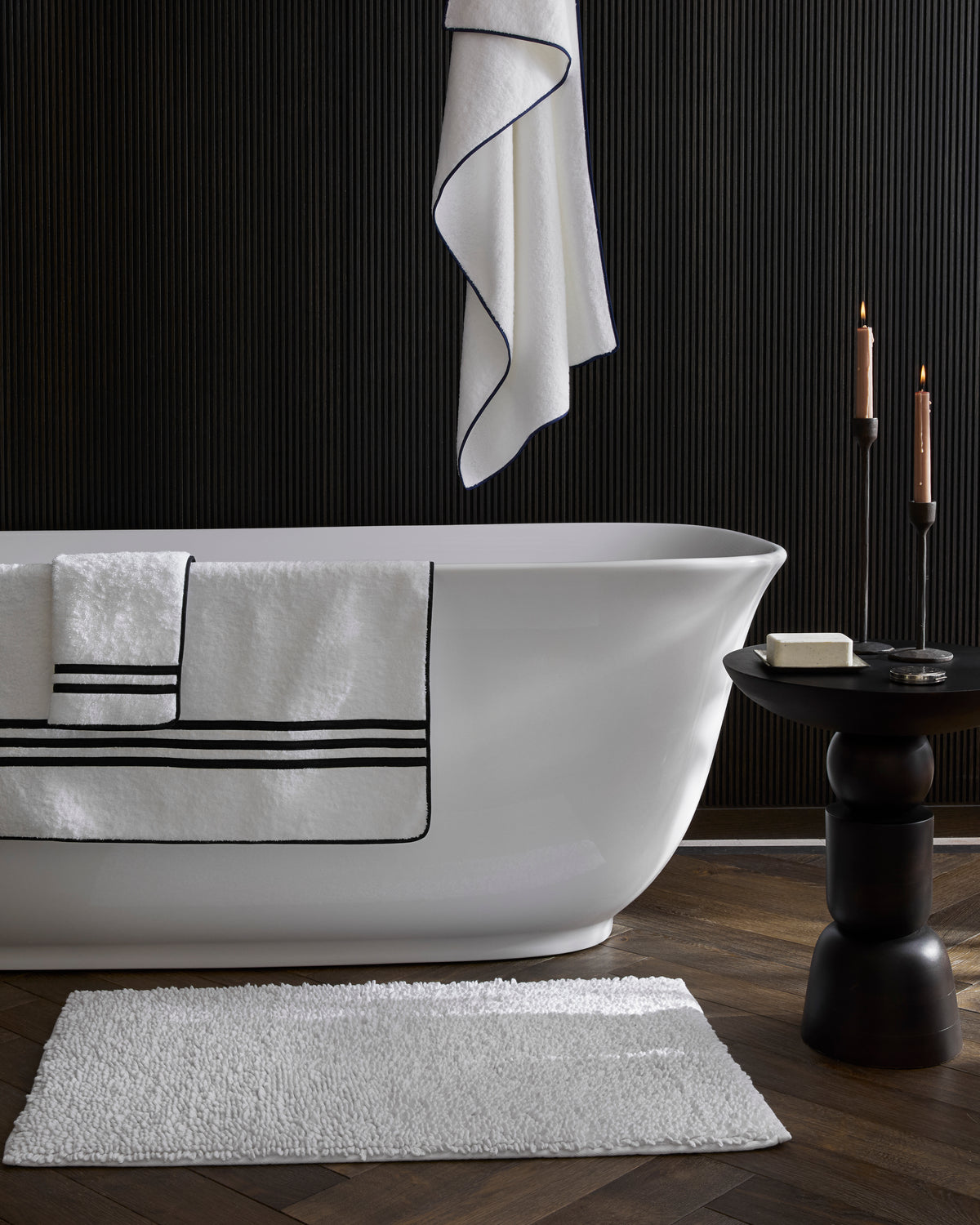 White chunky looped bath rug on wood floor in a modern bathroom with freestanding white tub and black paneled wall