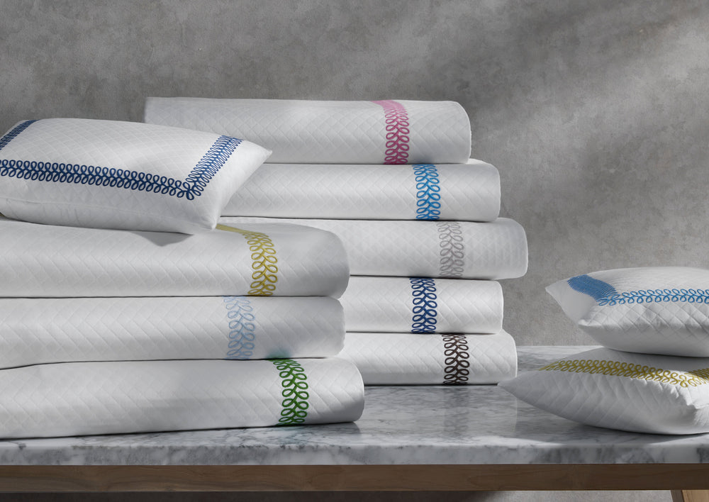 Stacked Astor Braid Matelassé coverlets and shams with colored braided trim and diamond quilting on a marble tabletop
