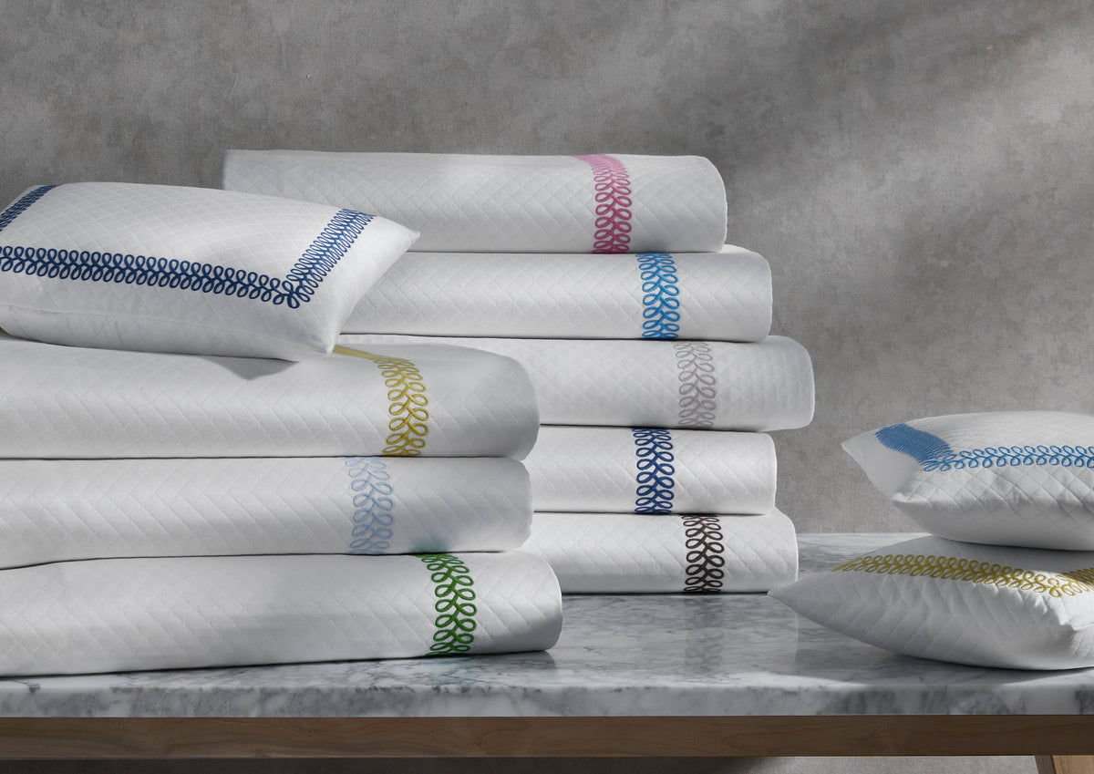 Stacked Astor Braid Matelassé coverlets and shams with colored braided trim and diamond quilting on a marble tabletop
