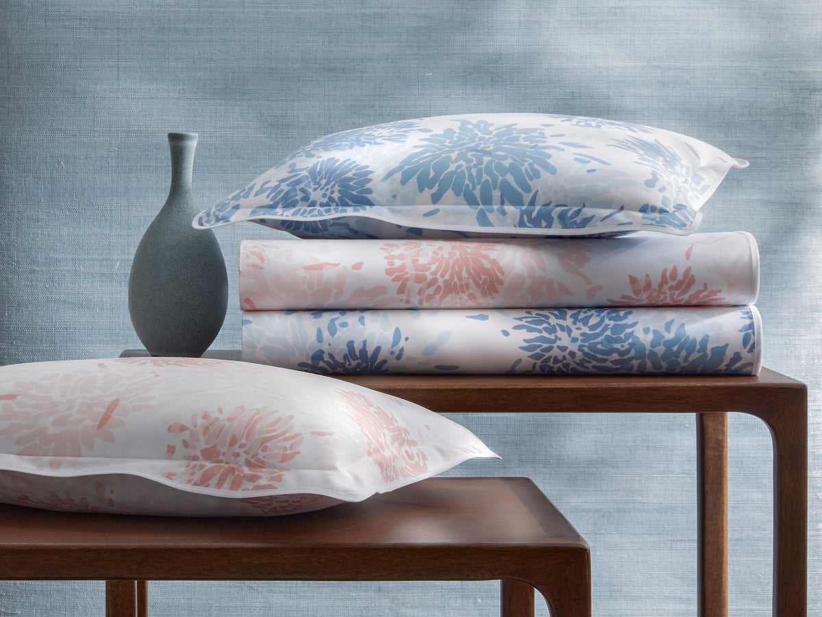 Stacked Chrysanthemum sham and folded sheets showing blue and blush floral print on white with blue textured backdrop