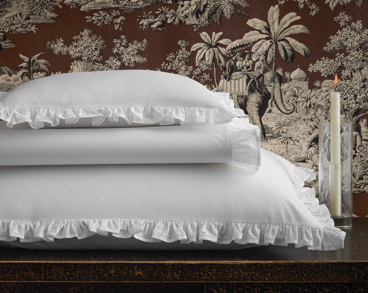 Stacked view of Georgia flat sheet and matching pillowcases showcasing white ruffled hemstitch against patterned toile wallpaper