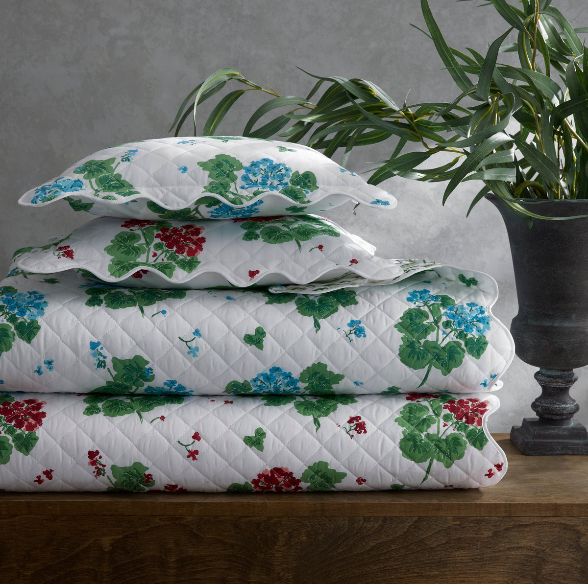 Stacked Geranium flat sheet and shams with large red and blue geranium print on white quilted fabric and scalloped edges on wood table