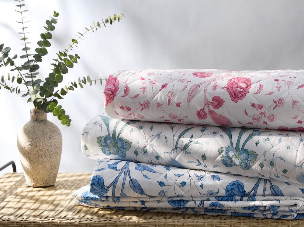 Stacked view of Khilana quilts showcasing pink, green, and blue floral prints on a wicker surface with a ceramic vase and eucalyptus