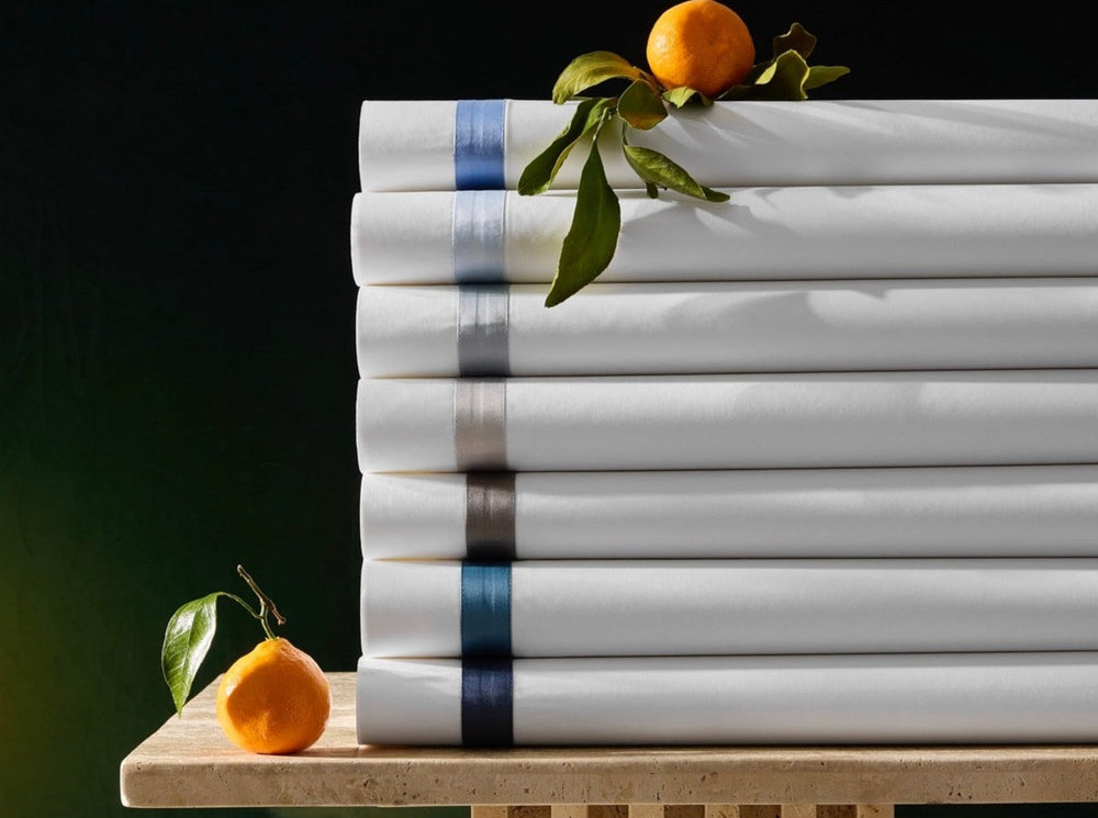 Stacked side view of Lowell bedding sheets showing colored ribbon hems on white fabric with tangerines on a stone table and dark green background