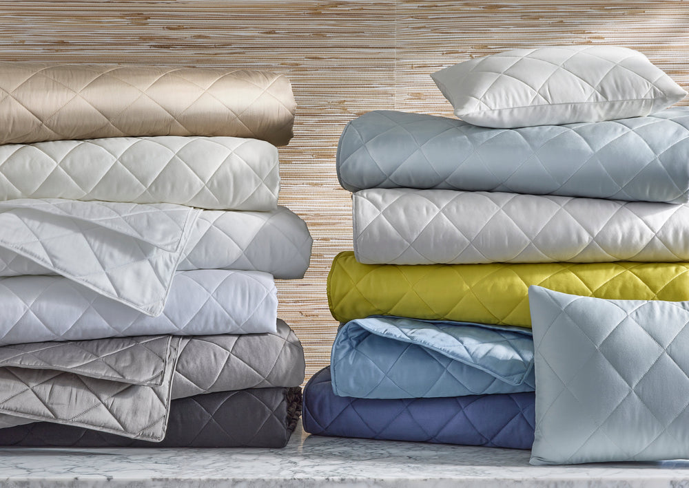 Front view of Nocturne quilts stacked to show diamond quilting and available colors (white, gray, blue, lemon) on a marble surface
