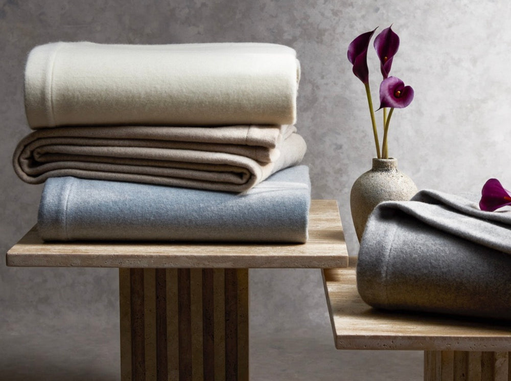 Three folded Venus blankets in ivory, dune, and pool stacked on a wooden pedestal with a rolled grey blanket and a small vase on a mottled grey backdrop