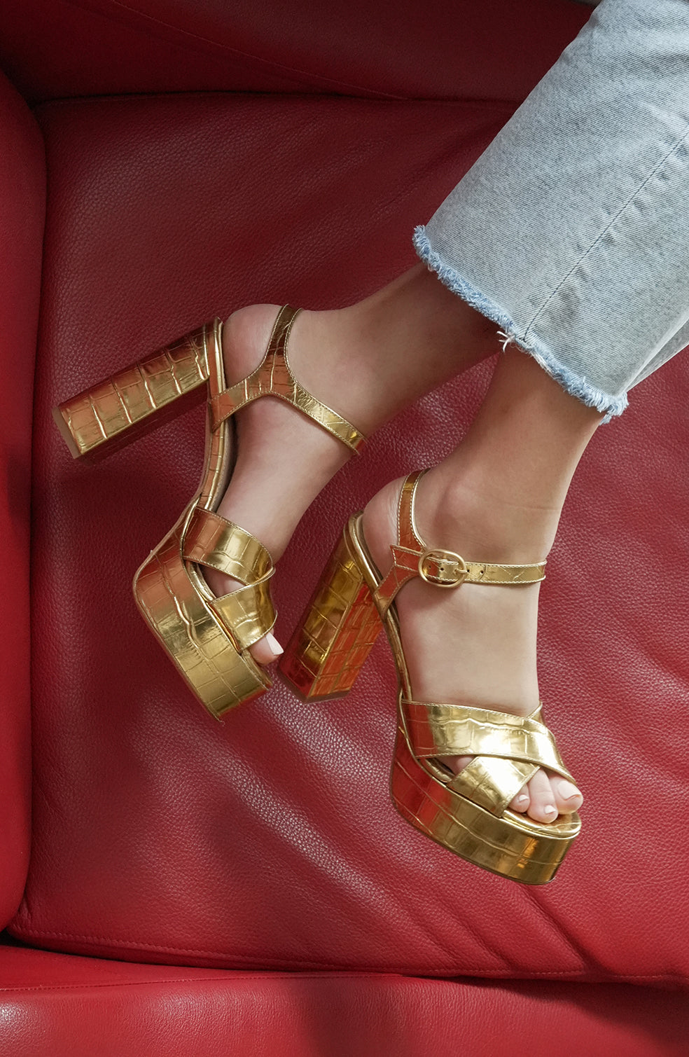 Angled view of Bella gold faux croc platform sandal with block heel, buckled ankle strap and croc texture on red background