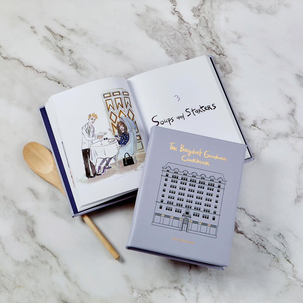 Angled view of The Bergdorf Goodman Cookbook in Lilac showing its illustrated hardcover and open pages on marble