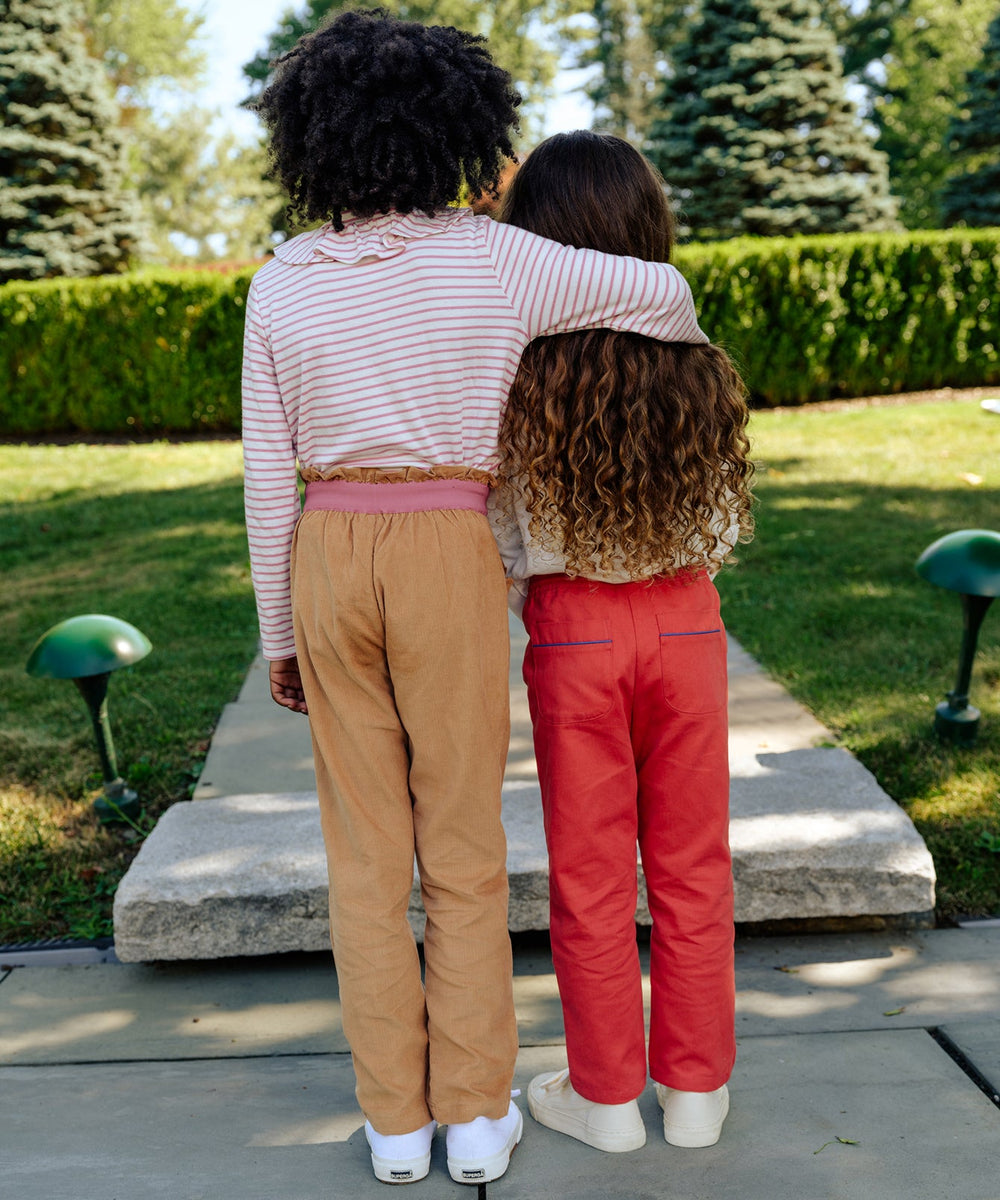 Two children standing arm-in-arm outdoors, one wearing honey corduroy Betsy pants with cuffable hem and ruffled elastic waist