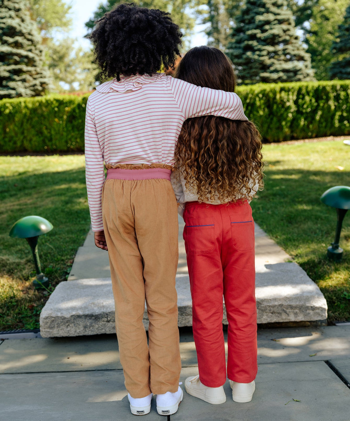 Two children standing arm-in-arm outdoors, one wearing honey corduroy Betsy pants with cuffable hem and ruffled elastic waist