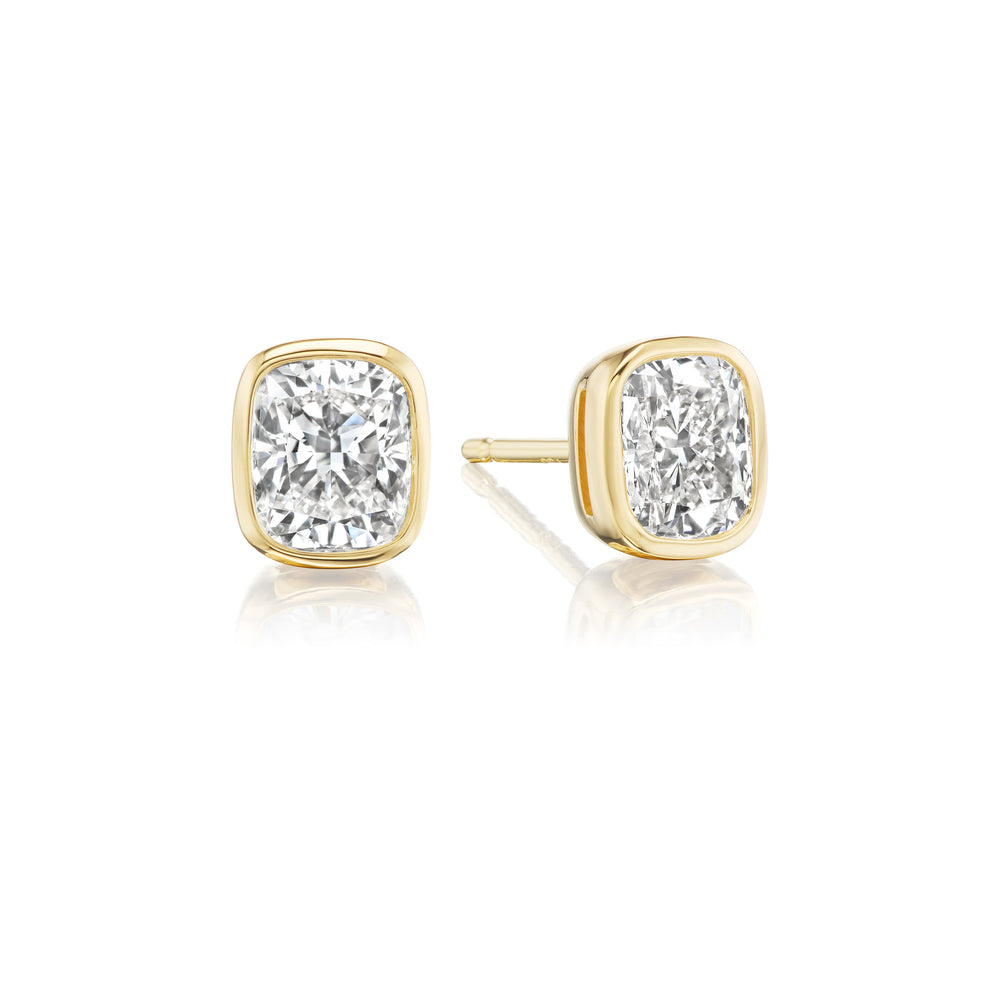 The Bezel Set Cushion Cut Diamond Studs in Yellow Gold feature VVS2 clarity diamonds, shown on a white background with one earring facing forward and the other displaying its side and post.