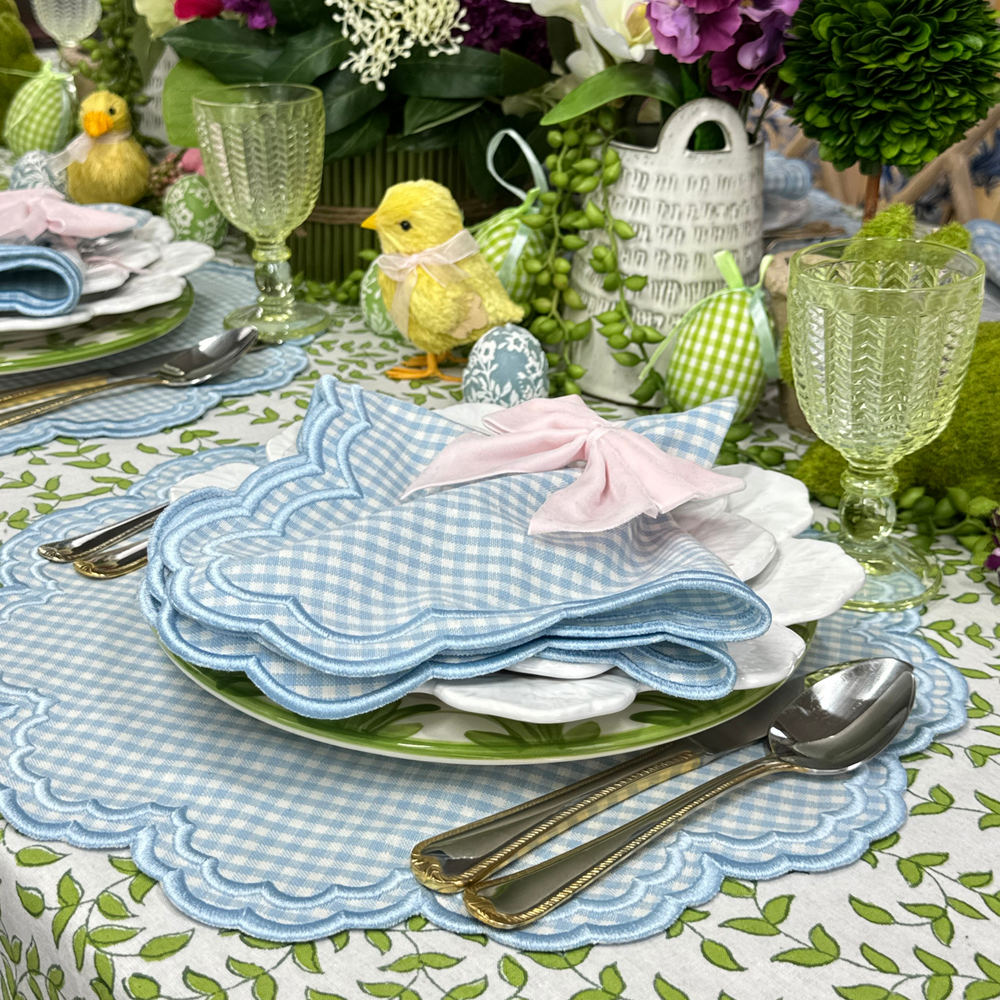 Bettina gingham napkins folded on stacked plates with a pink bow, showing blue gingham pattern and scalloped white embroidery on a floral table setting