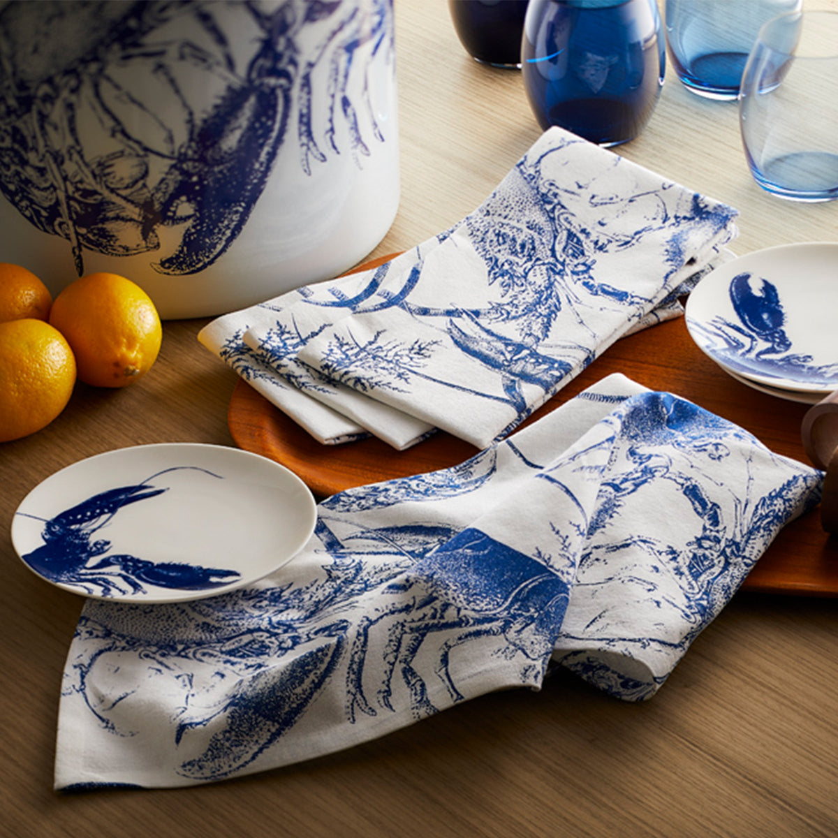 Blue lobster dinner napkins arranged on a table with matching plates, a pitcher, blue glasses, and oranges