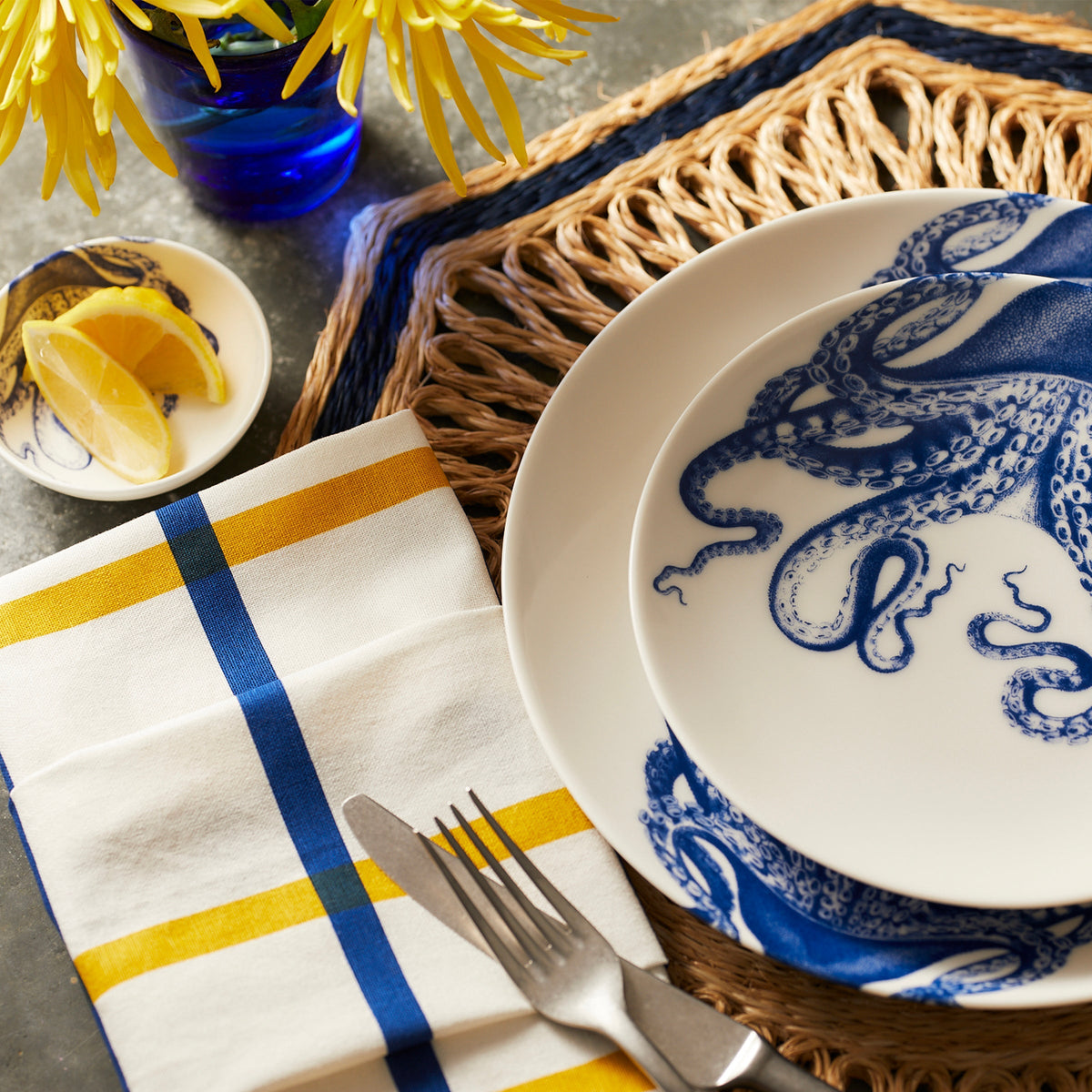 Blue and yellow plaid dinner napkin displayed on a table with patterned plates, cutlery, flowers, and woven placemat