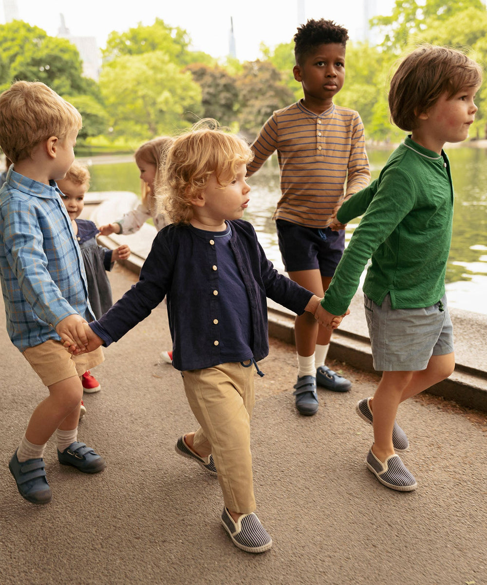 Group of toddlers holding hands and walking along a park pond, one child wearing khaki baby pants