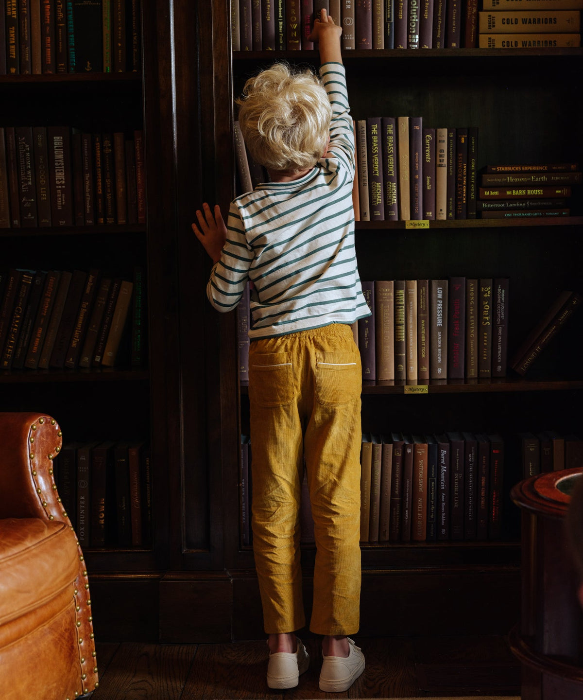 Back view of Bowie Pant in Mustard Corduroy showcasing cuffed bottoms and contrast pocket piping on a child in front of bookshelves