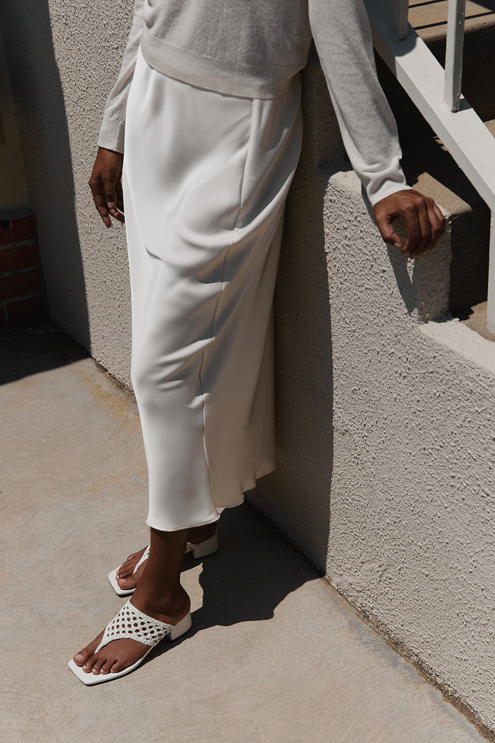 Close-up side view of Brenda off-white sandal showing woven thong upper, low block heel, and square toe on a model against sunlit concrete steps