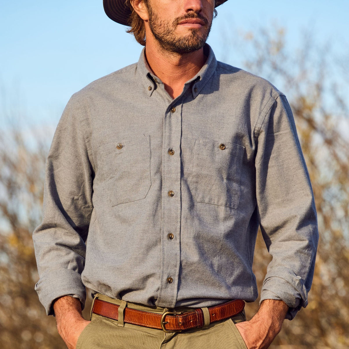 Man wearing a Tom Beckbe Dixon Brushed Cotton Twill Shirt, brown belt, and hat outdoors with trees in the background.
