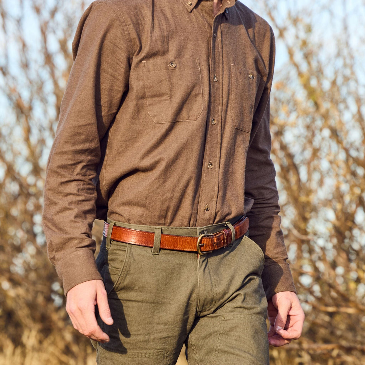 Wearing a Dixon Brushed Cotton Twill Shirt by Tom Beckbe, green pants, and a brown belt, someone walks near leafless bushes.